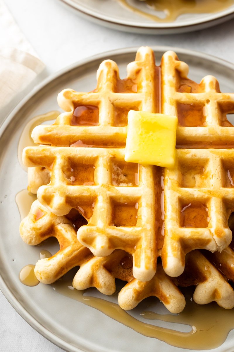 Gros plan d'une pile de gaufres sur une assiette beige, arrosées de sirop et garnies d'un bloc de beurre